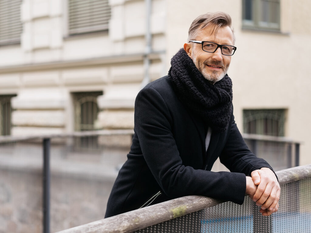 Mężczyzna w czarnym płaszczu i szaliku stoi na balustradzie w miejskim otoczeniu.