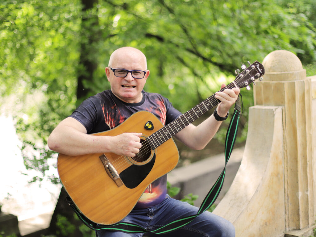 Mężczyzna grający na gitarze akustycznej w zielonym parku, uśmiechnięty i zaangażowany w występ.