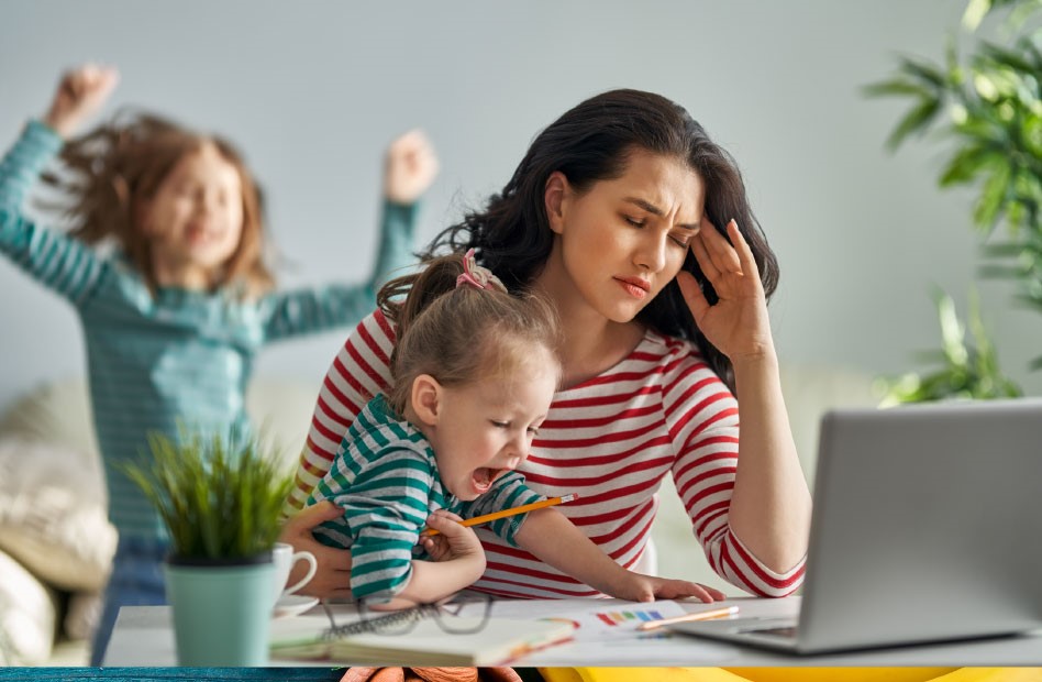 Zmienna mama z dzieckiem na kolanach i pracującą przy biurku zmaga się z przepracowaniem w domowym biurze.