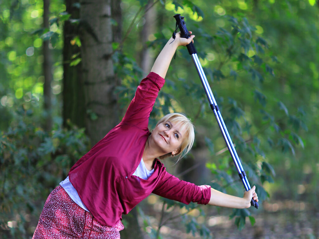 Kobieta ćwiczy rozciąganie w lesie z użyciem kijków do Nordic Walking, promując aktywny styl życia na świeżym powietrzu.