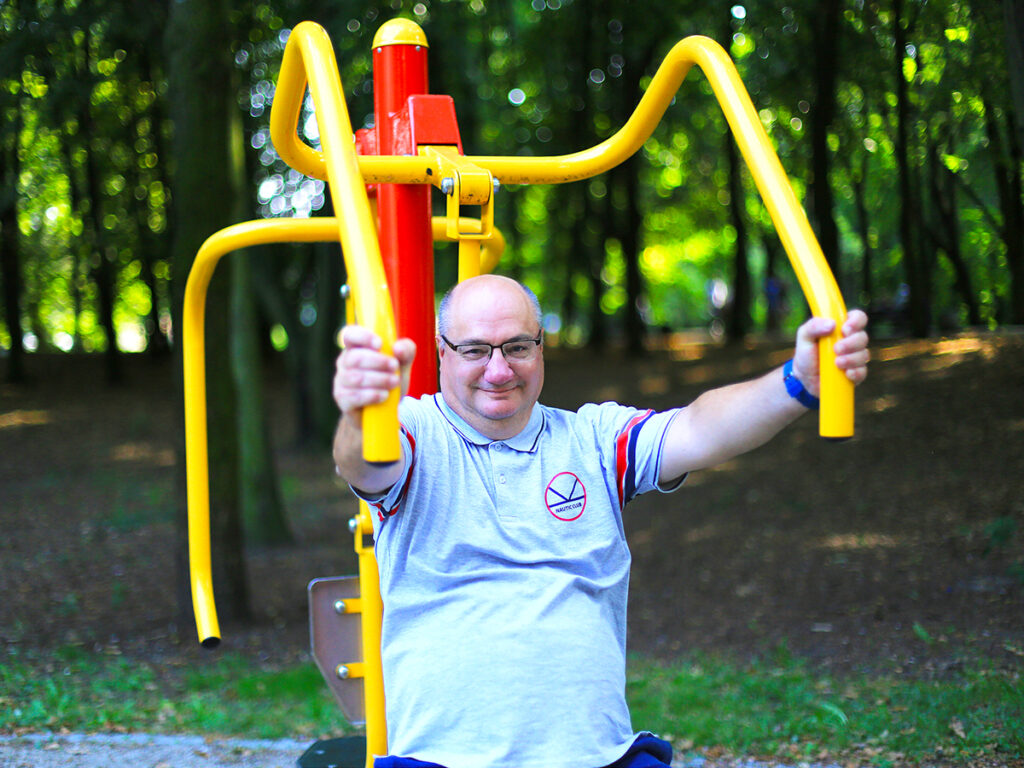 Mężczyzna ćwiczy na sprzęcie do fitnessu na świeżym powietrzu, otoczony zielenią drzew w parku.