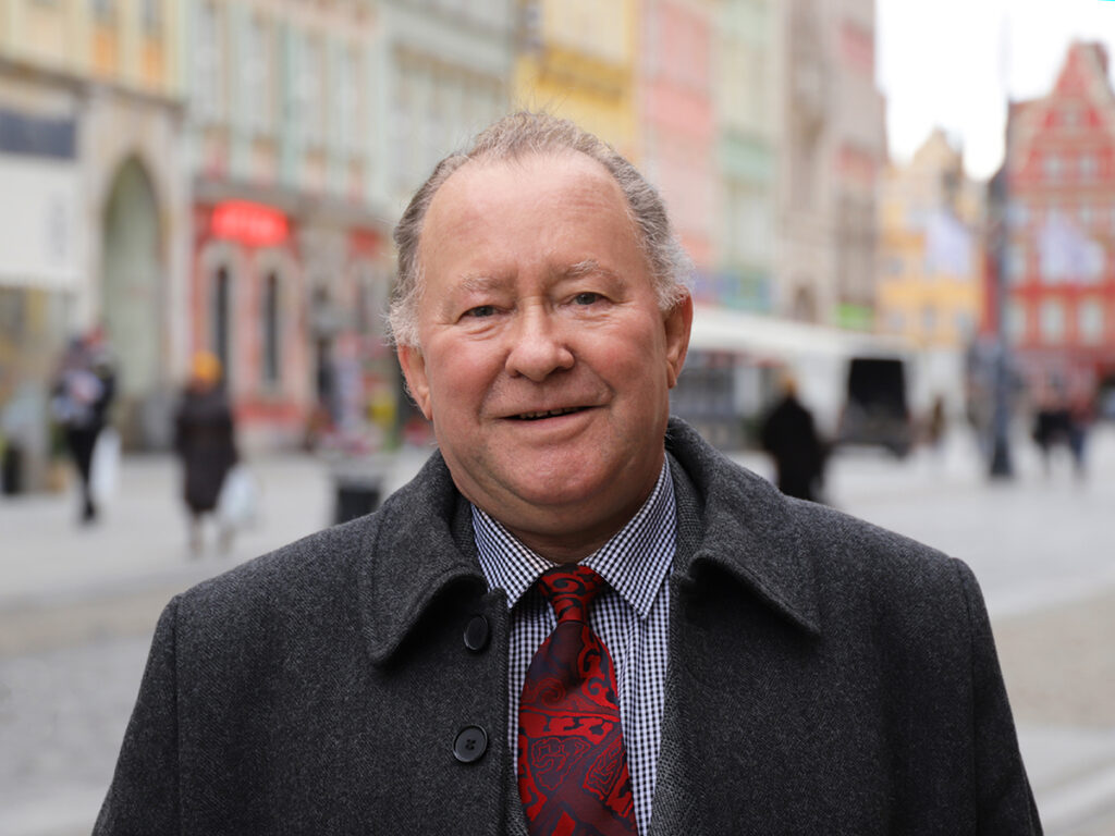 Mężczyzna w eleganckim płaszczu i krawacie stoi na ulicy, w tle kolorowe kamienice i przechodnie.