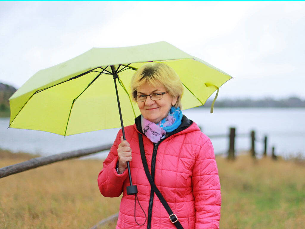 Kobieta w różowej kurtce trzyma jasnożółty parasol na tle jeziora w wietrznym otoczeniu.