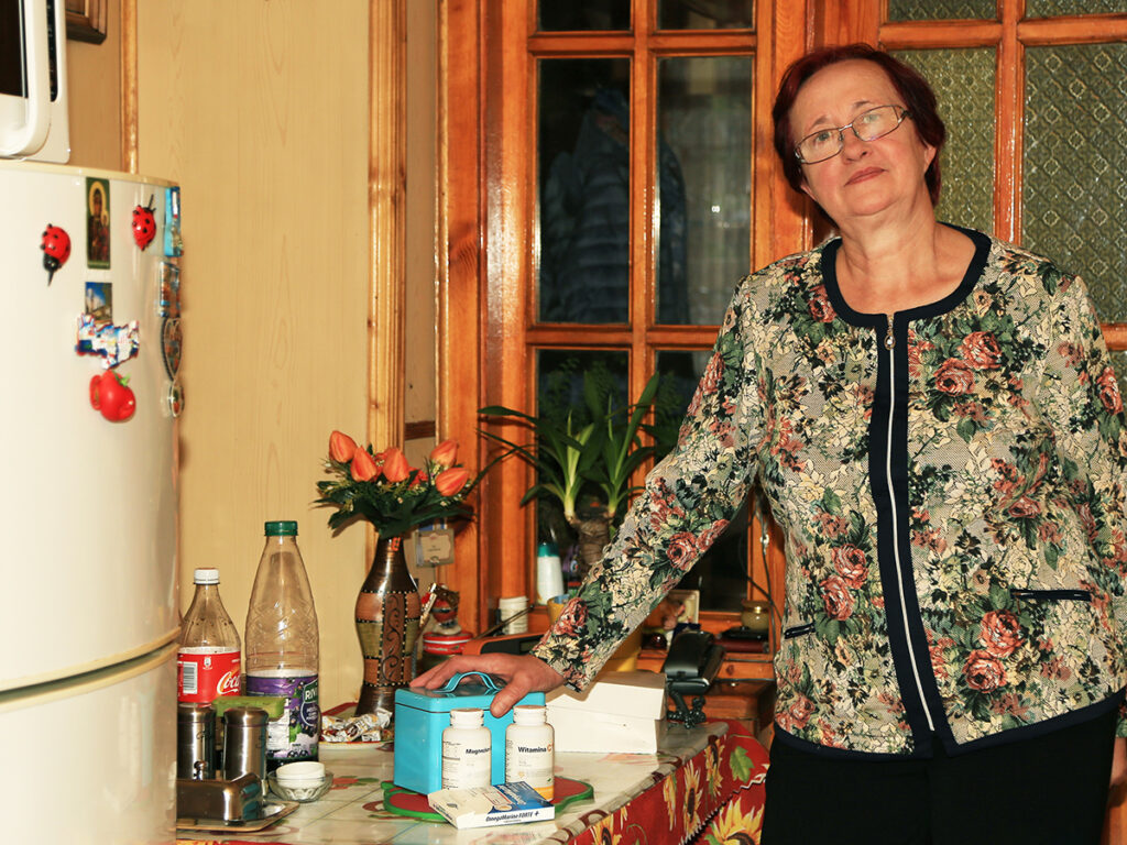 Kobieta w florystycznym swetrze stoi przy stole w kuchni, obok leków i butelek, w otoczeniu roślin i kwiatów.