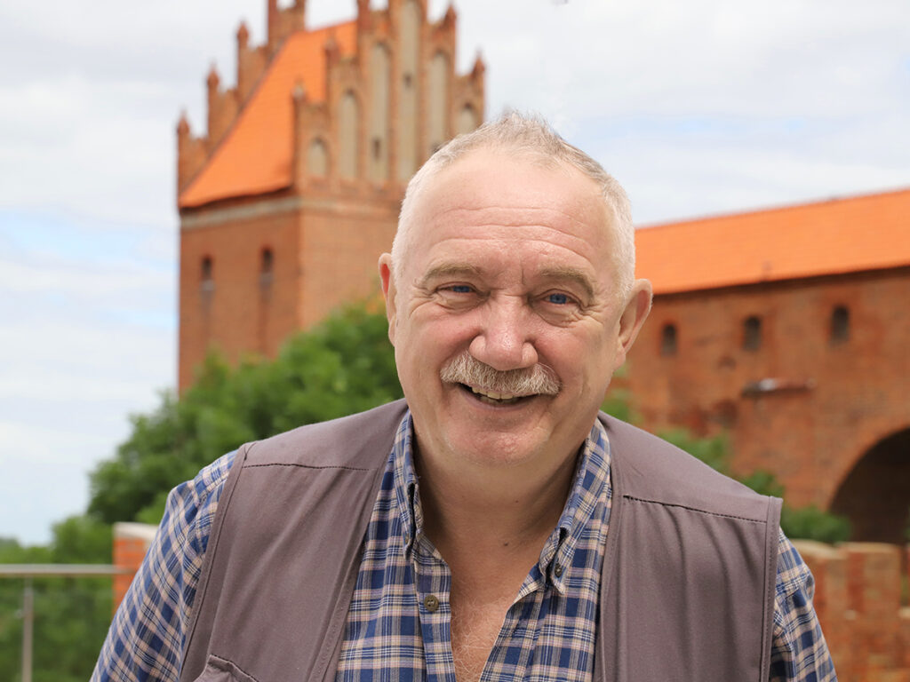 Mężczyzna o dużym uśmiechu stoi przed średniowiecznym zamkiem z pomarańczowym dachem, otoczony zielenią.