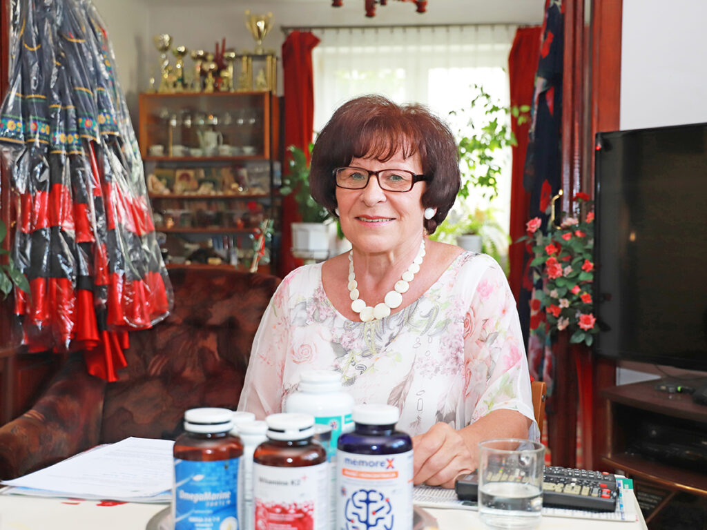 Starsza kobieta siedząca przy stole w przytulnym wnętrzu, otoczona suplementami diety i dokumentami, uśmiecha się do kamery.