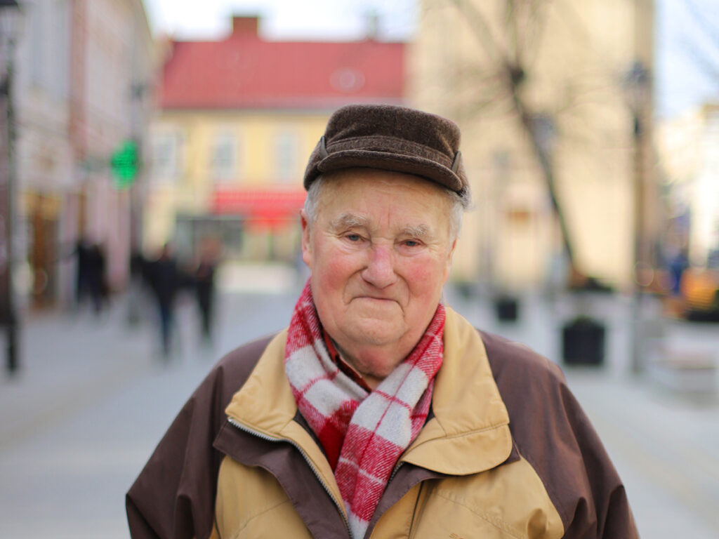Zdjęcie starszego mężczyzny w ciepłym płaszczu i czapce, uśmiechającego się na tle miejskiego krajobrazu.