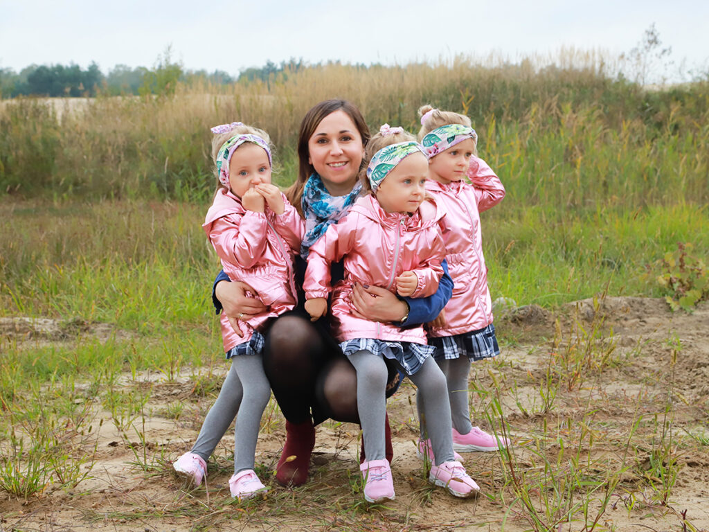 Na zdjęciu młoda mama w różowej kurtce trzyma na rękach dwie małe dziewczynki w identycznych różowych kurtkach, a trzecia stoi obok nich na tle zielonego krajobrazu.