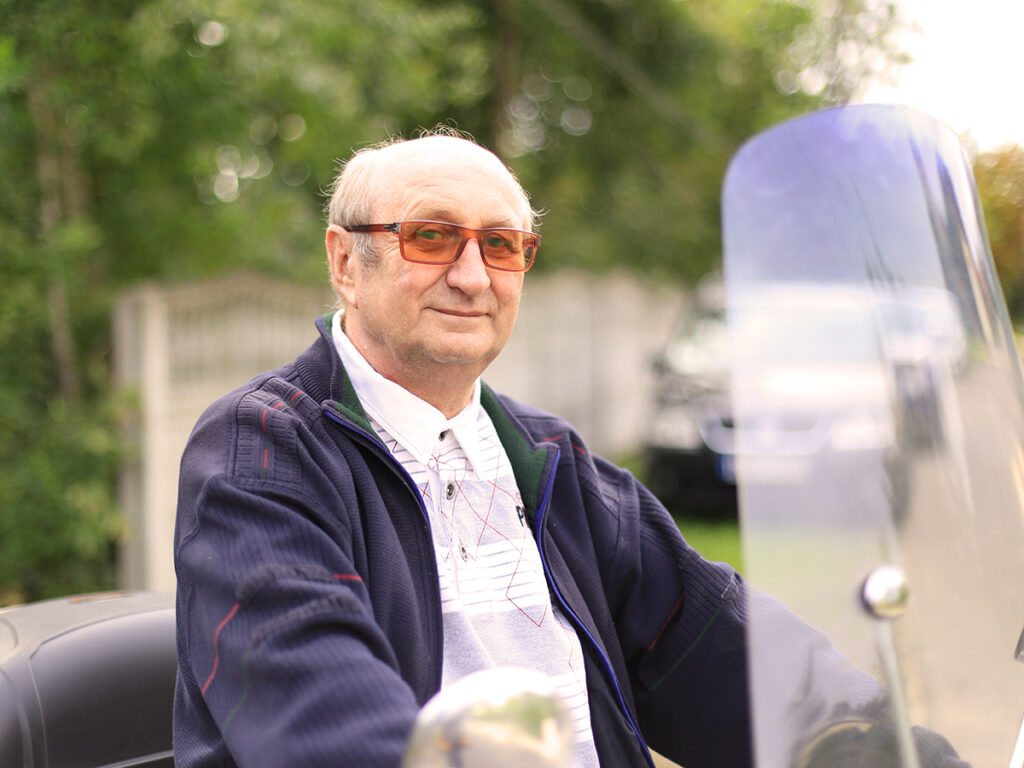 Starszy mężczyzna w okularach przeciwsłonecznych siedzi na motorze z wysuniętą przednią szybą, ciesząc się chwilą na świeżym powietrzu w otoczeniu zieleni.