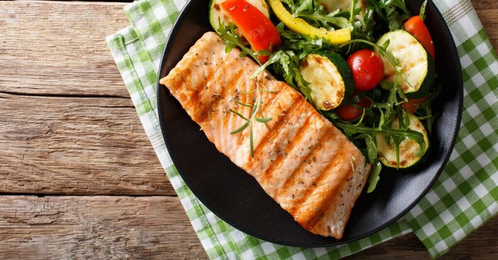 Grillowany łosoś podawany z sałatką z rukoli, pomidorów cherry i grillowanych cukinii na czarnym talerzu.