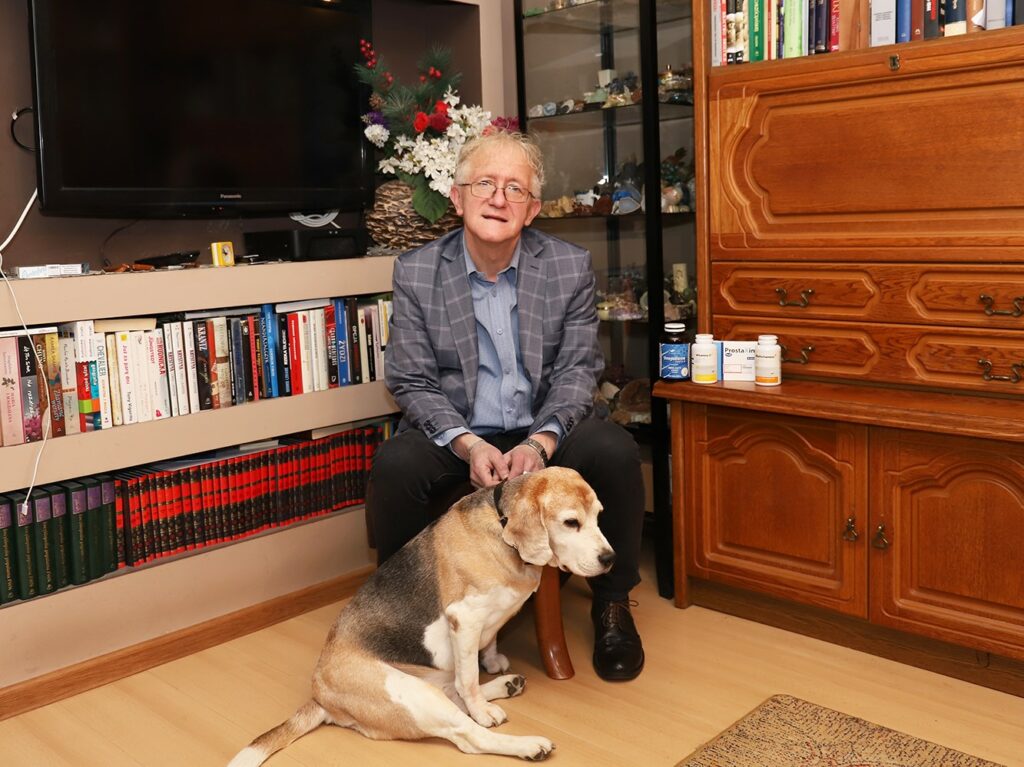 Mężczyzna w okularach w eleganckiej marynarce siedzi na podłodze w domowym wnętrzu obok beagle'a, otoczony książkami oraz dekoracjami.