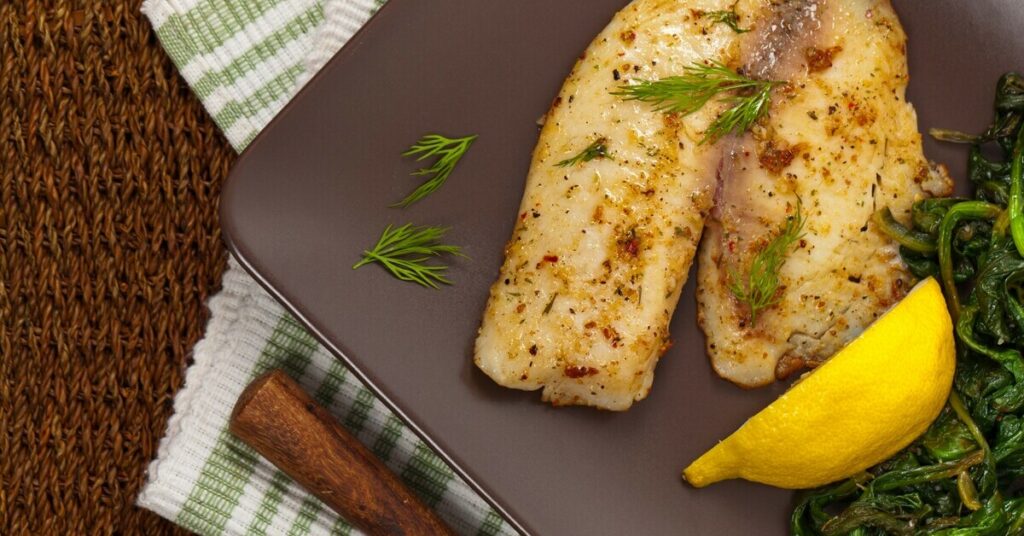 Grillowana ryba z koperkiem, podana z cytryną i duszonymi szpinakiem, jako zdrowe danie na lekki obiad.