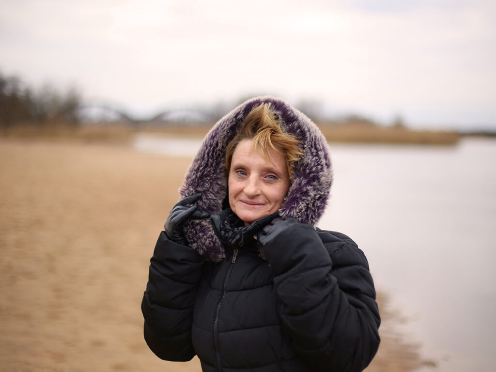 Kobieta w ciepłej kurtce z kapturem na tle plaży i rzeki, uśmiechająca się w chłodny, pochmurny dzień.