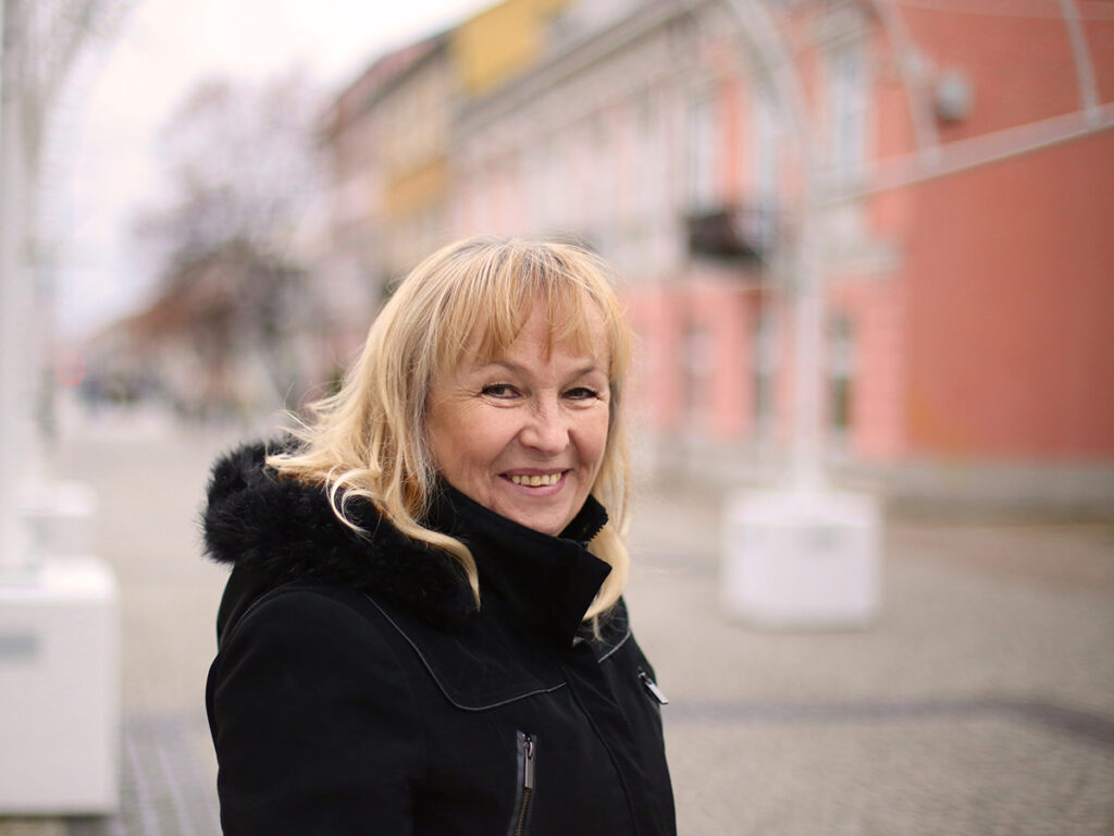 Uśmiechnięta kobieta z blond włosami stoi na ulicy, w tle widać kolorowe budynki.