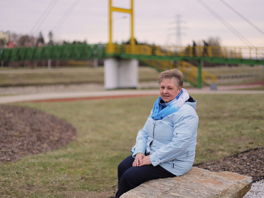 Starsza kobieta w niebieskiej kurtce i szaliku siedzi na głazie w parku, z mostem w tle.