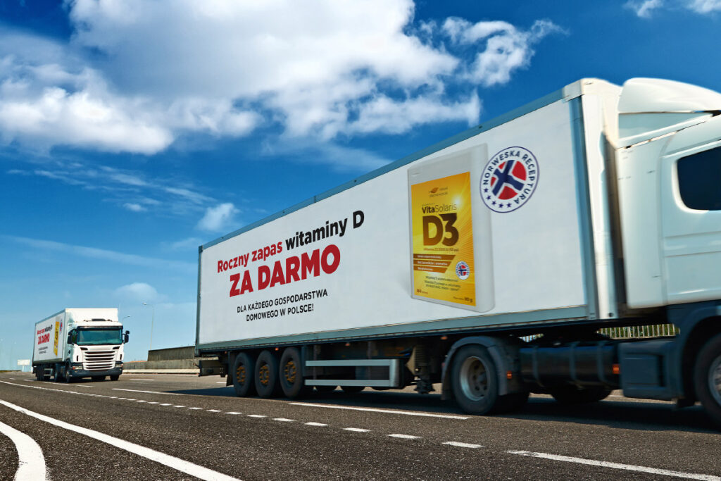 Na zdjęciu widoczny jest przejeżdżający ciężarówka z reklamą suplementu witaminy D3, promującą roczny zapas dostępny za darmo dla gospodarstw domowych w Polsce.