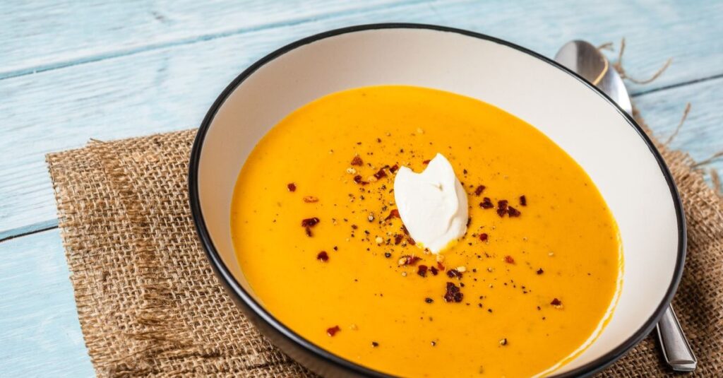 Zupa dyniowa w eleganckiej misce, udekorowana bitą śmietaną i przyprawami, na tle juty i niebieskiego blatu.