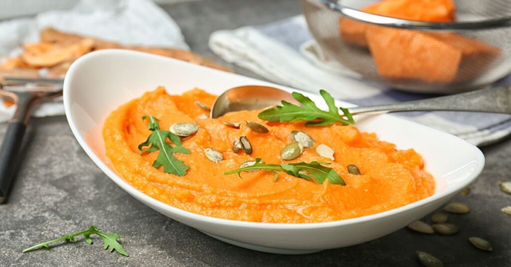 Puree z batata w białej misce, ozdobione nasionami i świeżą zieleniną, na tle kuchennego blatu.