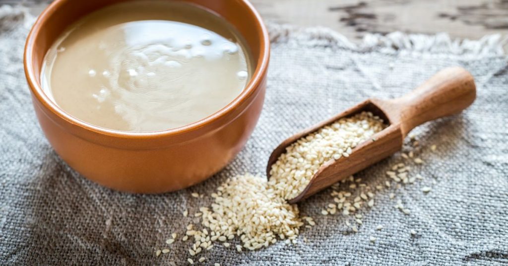 Miska z pastą tahini obok łyżki sezamu na lnianym tle, idealna do zdrowych przepisów.