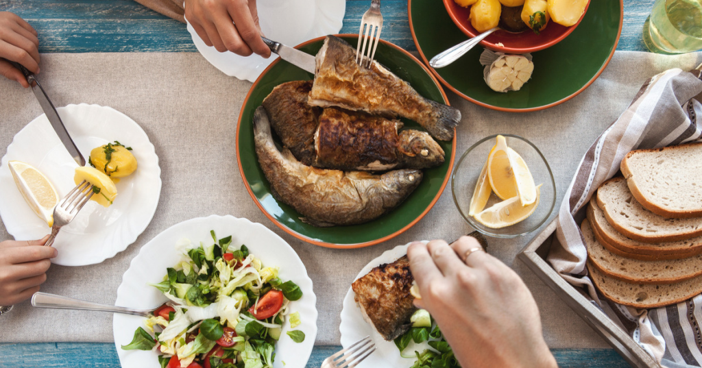 Stół zastawiony pysznymi potrawami, w tym grillowanymi rybami, świeżą sałatą, cytrynami, ziemniakami oraz chlebem, zachęca do wspólnego posiłku.