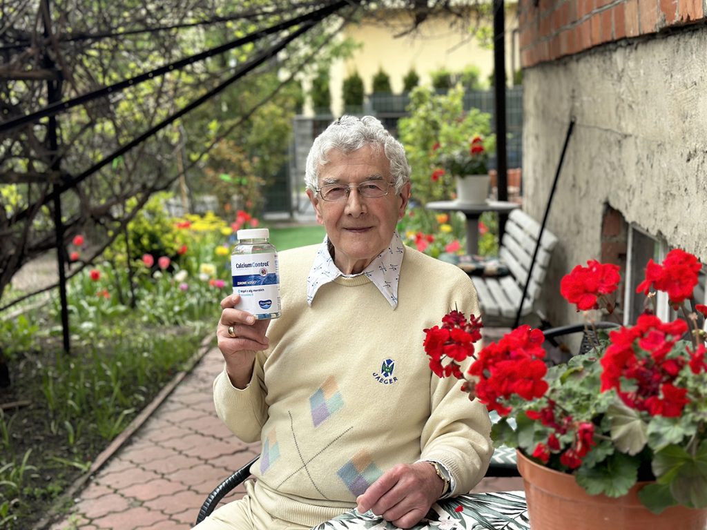 Starszy mężczyzna w ogrodzie trzyma suplement diety Calcium Control, promując zdrowy styl życia i dbanie o kości.