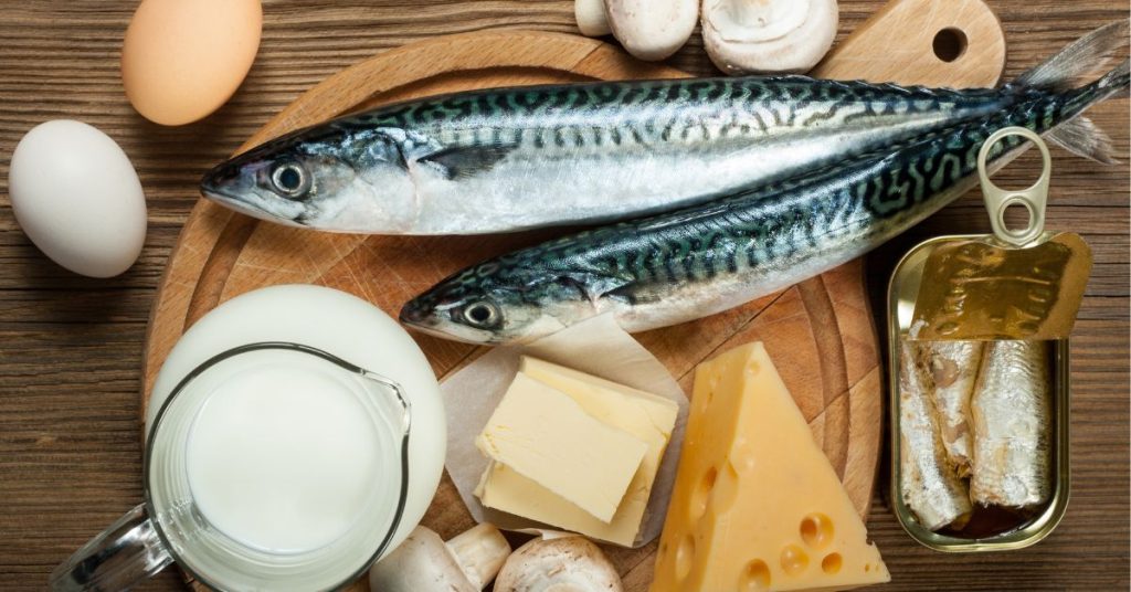 Na drewnianej desce znajduje się zestaw składników bogatych w białko, w tym świeża makrela, jaja, ser, masło oraz mleko, idealnych do przygotowania zdrowych posiłków.