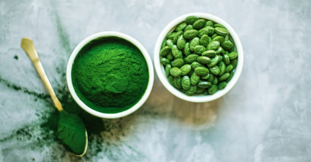 Obraz przedstawia miski z zielonym proszkiem i nasionami spiruliny, które są popularnymi suplementami zdrowotnymi bogatymi w składniki odżywcze.
