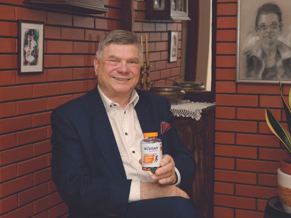 Mężczyzna w eleganckim ubraniu trzyma produkt o nazwie Movisan, uśmiechając się w przytulnym wnętrzu z ceglanymi ścianami.