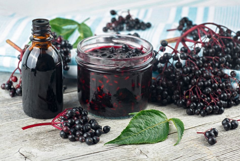 Zdjęcie przedstawia ciemny syrop z czarnego bzu oraz butelkę, otoczone świeżymi owocami i liśćmi tej rośliny na drewnianym tle.