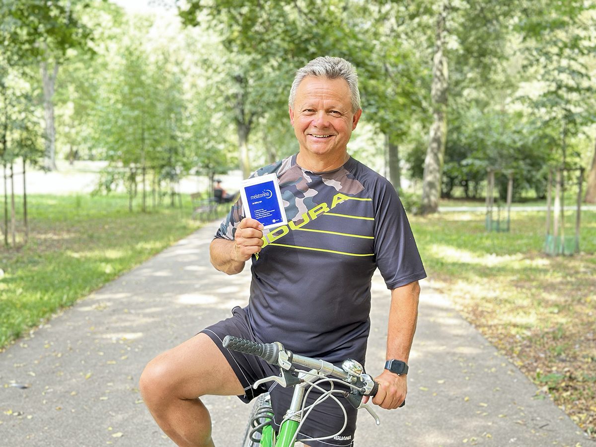 Mężczyzna w sportowej koszulce stoi na rowerze w parku, trzymając w ręku suplement diety Melatonina