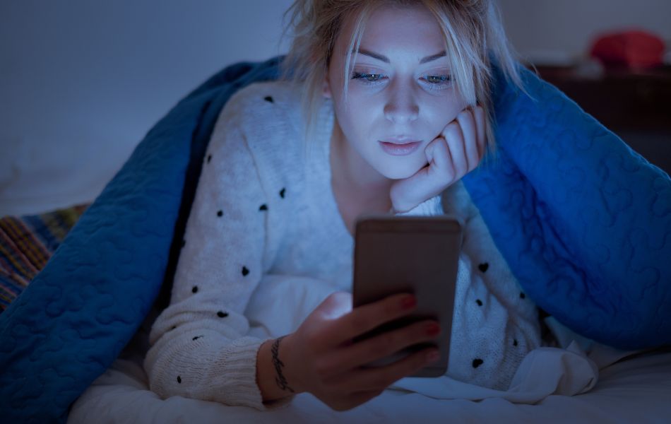 Młoda kobieta w swetrze leży w łóżku pod niebieskim kocem, skupiona na ekranie telefonu w przytulnej, wieczornej atmosferze.
