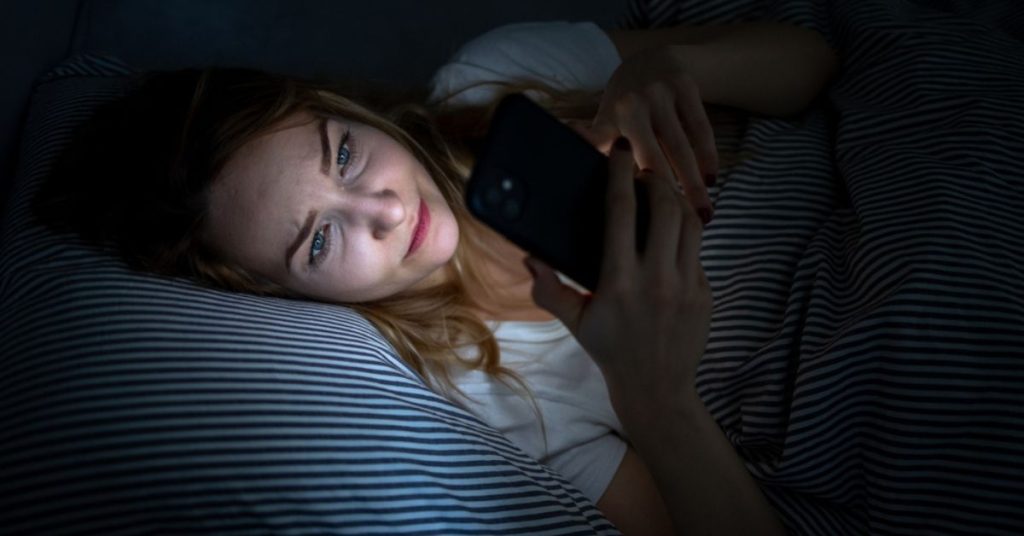 Młoda kobieta leży na poduszce, patrząc na telefon komórkowy w ciemnym pokoju, co ilustruje wpływ technologii na sen.