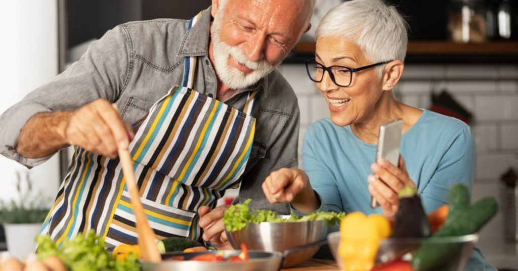 Dwoje seniorów radosne gotuje w kuchni, otoczeni świeżymi warzywami i używając nowoczesnych narzędzi kuchennych.