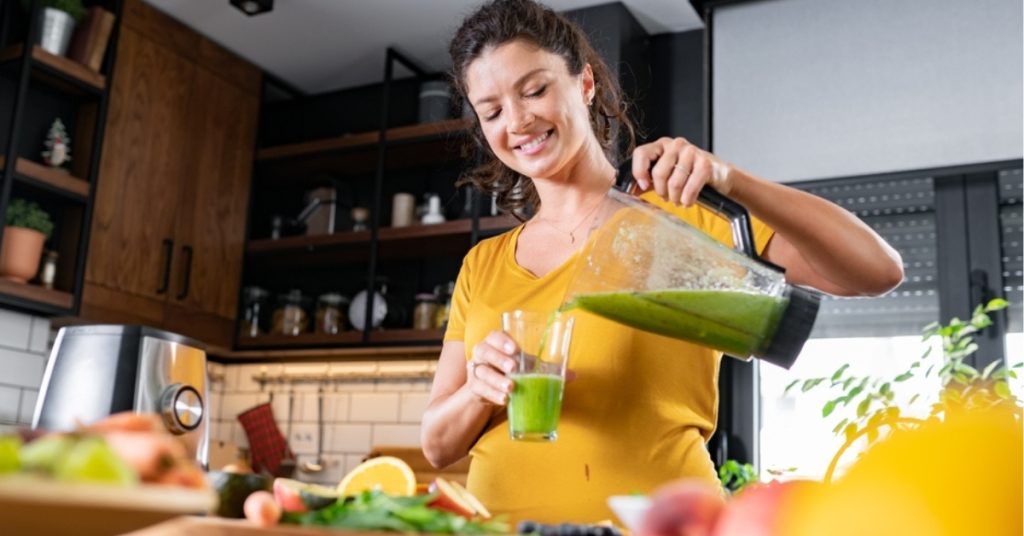 Kobieta przygotowująca zdrowy smoothie w nowoczesnej kuchni, otoczona świeżymi owocami i warzywami.