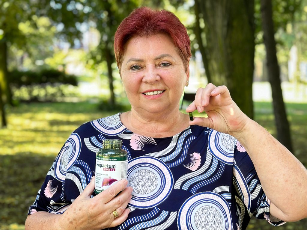 Kobieta trzymającą w dłoni kapsułkę suplementu diety Hepactum Forte, uśmiecha się na tle zielonego parku.