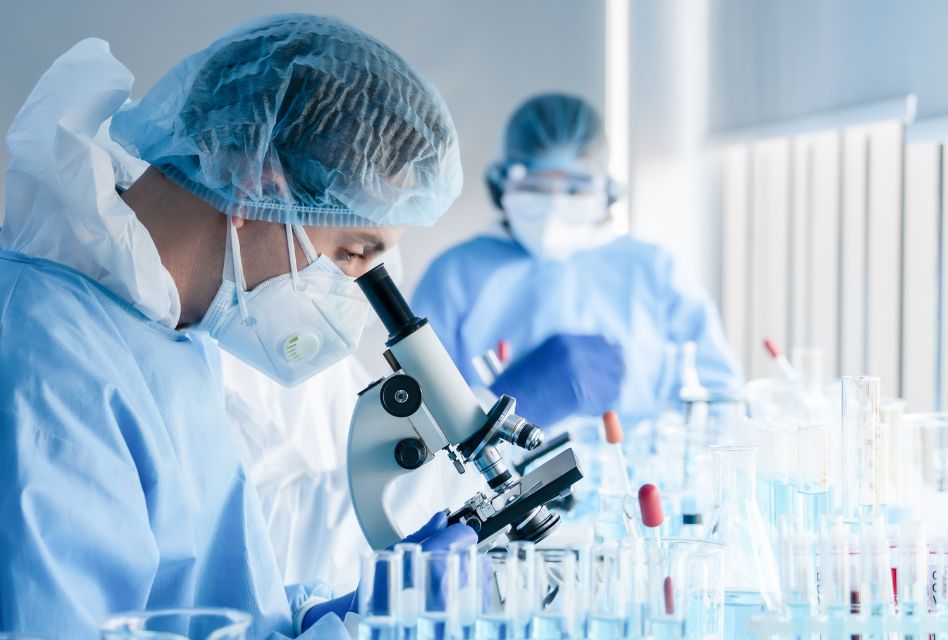 Laboratorium naukowe z dwóch pracowników w odzieży ochronnej, którzy prowadzą badania pod mikroskopem. Na stole znajdują się probówki z niebieskimi cieczami, co wskazuje na wykonywanie analizy chemicznej. Scena ukazuje profesjonalne podejście do pracy w biotechnologii lub chemii, z dbałością o higienę i bezpieczeństwo.