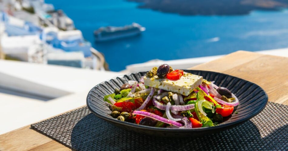 Sałatka grecka z serem feta, oliwkami, pomidorkami i czerwoną cebulą, podana na eleganckim czarnym talerzu. Tło obrazu przedstawia malowniczy widok na morze oraz architekturę Santorini, co podkreśla letni klimat i urok tej potrawy. Idealna na zdrowy obiad lub kolację w słoneczny dzień.