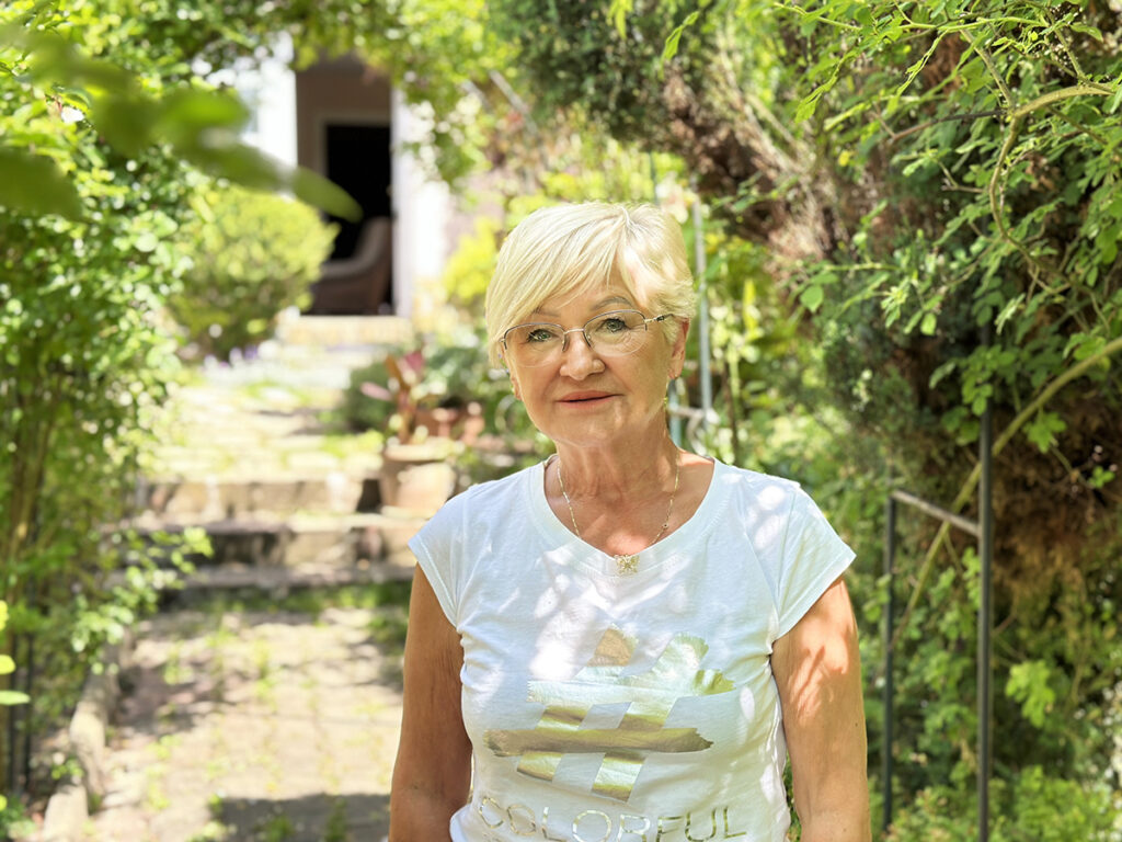 Kobieta z jasnymi włosami i okularami stoi w bujnym ogrodzie, uśmiechając się do aparatu.